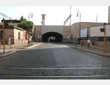 Strada urbana che conduce a un sottopasso ad arco in muratura, affiancato da edifici bassi con graffiti. Ai lati ci sono marciapiedi con lampioni, segnali stradali e cartelloni, con un incrocio in primo piano.