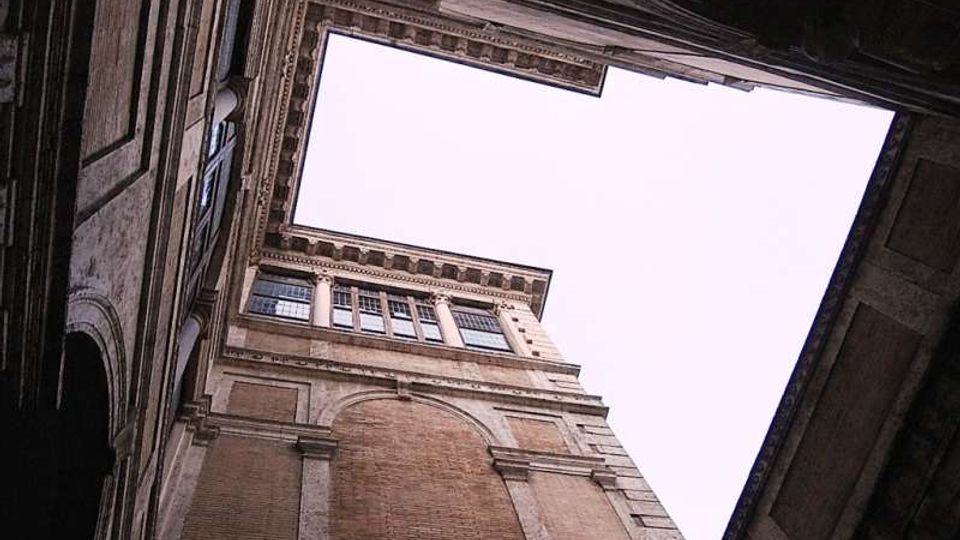 Veduta dal basso di un cortile interno con facciate alte in muratura e pietra, caratterizzate da cornici decorative, finestre rettangolari e un grande riquadro arcuato. Il cielo è visibile attraverso un’apertura superiore irregolare delimitata dai cornicioni del palazzo.