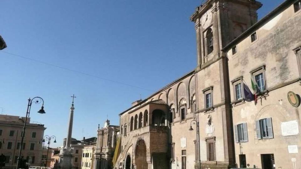 Piazza cittadina con grande edificio storico in pietra e mattoni, con archi, finestre e una torre campanaria laterale, affacciato sulla strada. Sullo sfondo si vede un obelisco con croce e lampioni, con auto parcheggiate e altri palazzi lungo la piazza sotto cielo sereno.