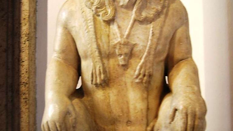 Scultura in pietra esposta su supporti in un ambiente museale, raffigurante una figura accovacciata con barba e capelli scolpiti. Sfondo con parete chiara e base espositiva scura.