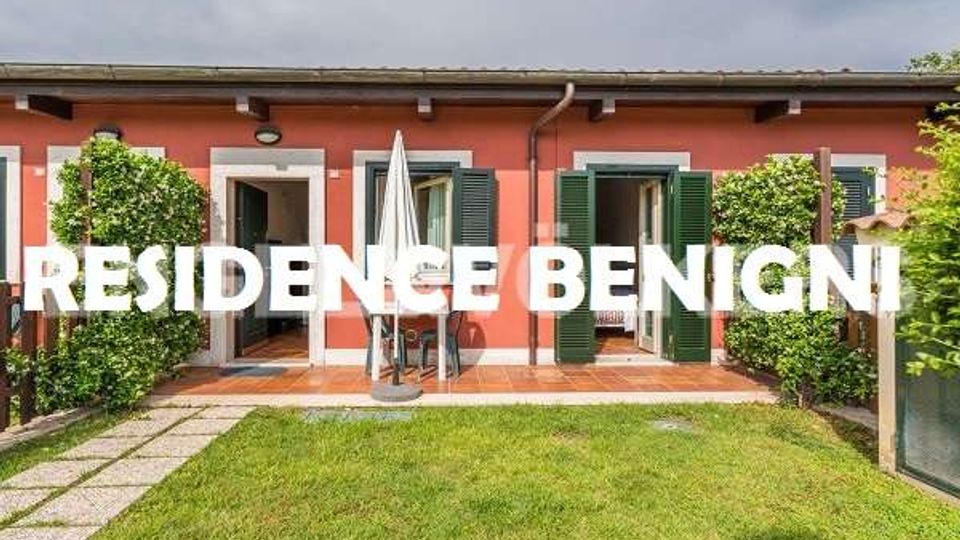 Facciata di una villetta a un piano color salmone con persiane verdi e porte-finestre aperte su un piccolo patio con tavolo e ombrellone. Davanti c’è un giardino con prato, siepi laterali e un camminamento in pietra.