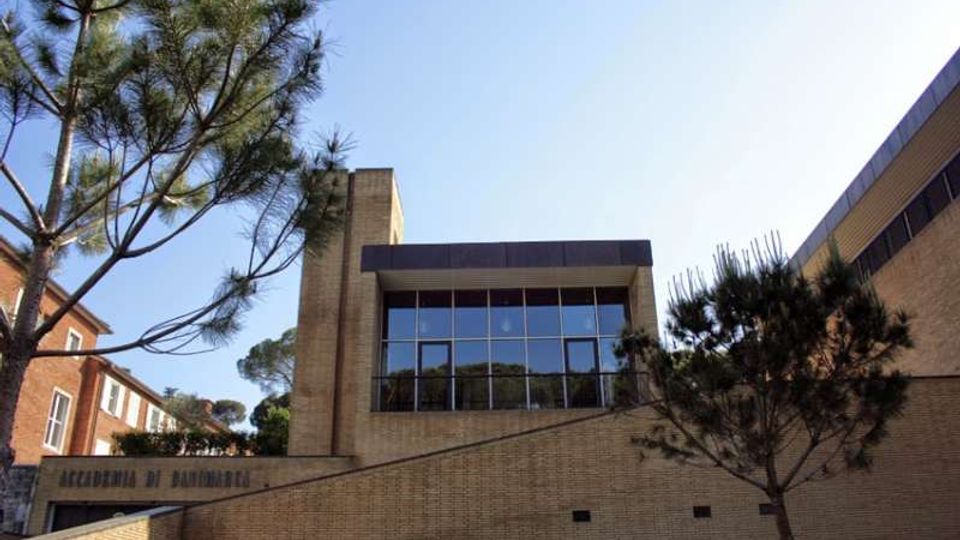 Edificio moderno in mattoni con ampie vetrate a griglia sulla facciata, affiancato da una struttura a torre e da un ingresso su un livello inferiore con muro in pendenza. L’area esterna è circondata da pini e altri edifici in mattoni sullo sfondo.