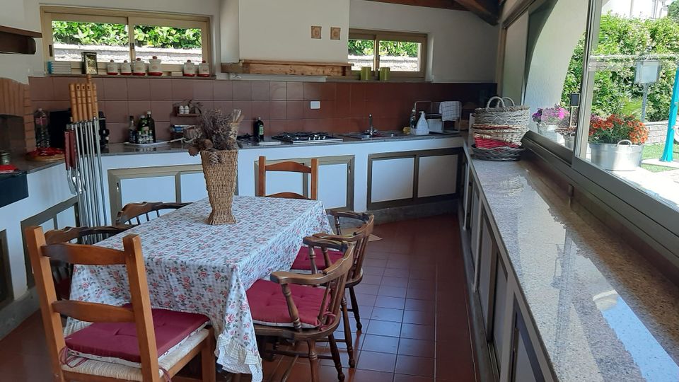 Cucina abitabile con soffitto in legno a vista, lunga penisola con piano chiaro e pareti piastrellate, affiancata da un tavolo con sedie in legno. Ampie finestre e una grande vetrata laterale illuminano l’ambiente e si affacciano sul giardino esterno.