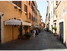 Strada stretta in ciottoli tra edifici alti dai colori ocra e arancio con persiane scure, con tende bianche e fioriere lungo il lato sinistro. La via prosegue in prospettiva con lampioni a parete, portoni e finestre sui fronti degli edifici.