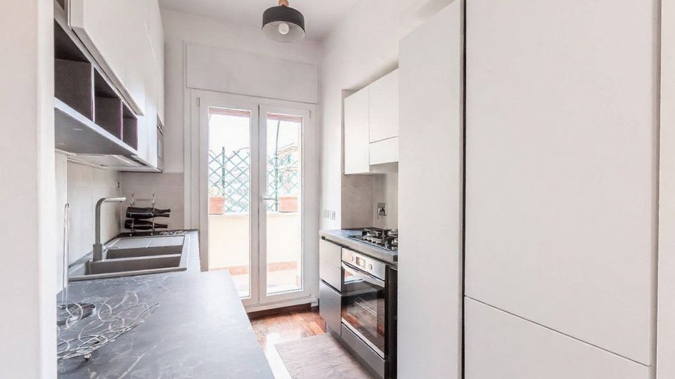 Cucina stretta e moderna con mobili bianchi e piani di lavoro grigi, dotata di lavello, forno e piano cottura lungo due pareti. In fondo, una portafinestra a doppia anta conduce a un piccolo balcone con grata e vasi.