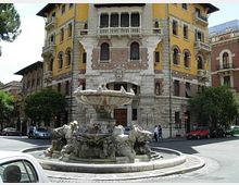 Fontana monumentale in pietra al centro di una rotonda stradale, con vasca circolare e sculture decorative alla base. Sullo sfondo si vede un grande edificio storico a più piani con facciata gialla e dettagli in pietra, archi e balconi, affacciato su un incrocio urbano.
