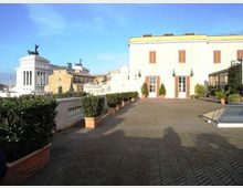Terrazza panoramica pavimentata in mattoni a spina di pesce con vasi di siepi lungo la balaustra, affacciata su edifici storici. Sullo sfondo si vede un palazzo bianco con persiane in legno e, a sinistra, un grande monumento neoclassico con statue sulla sommità.