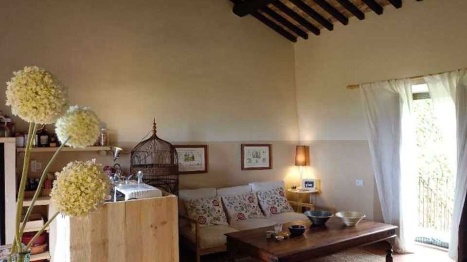 Soggiorno in stile rustico con soffitto alto e travi in legno a vista, divano con cuscini decorati e grande tavolino basso in legno. A sinistra si vede un angolo cucina con scaffali e bancone, mentre a destra una porta-finestra con tende bianche apre su un balcone.