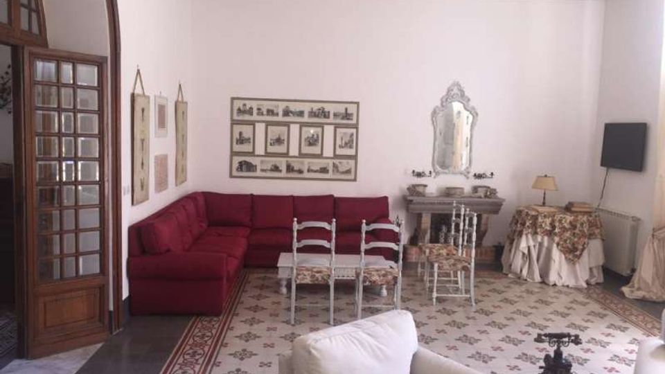 Salotto in stile classico con pavimento in piastrelle decorate, grande divano angolare bordeaux e sedie bianche attorno a un tavolino. Sulla parete si vedono stampe incorniciate e, a destra, un camino con specchiera ornata; a sinistra una porta ad arco in legno con vetri.