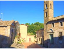 Piazzetta di un borgo con case in pietra e intonaco e una chiesa in muratura con campanile alto e aperture ad arco. Sullo sfondo si vedono colline alberate e cielo sereno, con scalinate e stradine lastricate che convergono nella piazza.