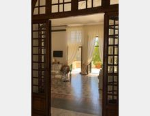 Interno di un salone luminoso visto attraverso una grande porta in legno e vetro, con pavimento in piastrelle decorate, tavolo con sedie e arredi classici. Alte tende chiare incorniciano porte-finestre aperte su un balcone con balaustra e grandi vasi di piante.
