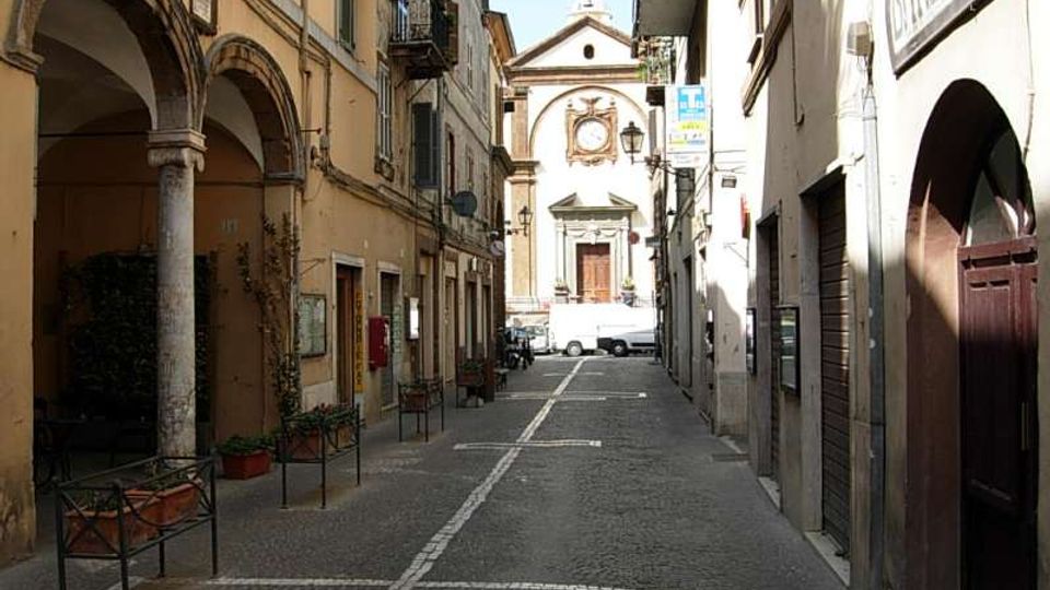 Via stretta in un centro storico con pavimentazione in pietra, edifici alti ai lati e portici ad archi con colonne sul lato sinistro. In fondo alla strada si vede la facciata di una chiesa con timpano e rosone, con un veicolo parcheggiato davanti.