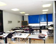 Aula scolastica con file di banchi e tavoli, soffitto con pannelli e luci rettangolari, e una lavagna sul fondo. Grandi finestre con tende blu lungo una parete illuminano lo spazio, con pareti bianche e pavimento in graniglia.