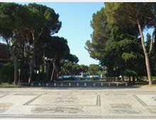 Ampio piazzale pavimentato con motivi decorativi, delimitato da una fila di dissuasori e circondato da alti pini e altre conifere. Sullo sfondo si intravede un’area verde con strutture e cartelli, sotto un cielo sereno.