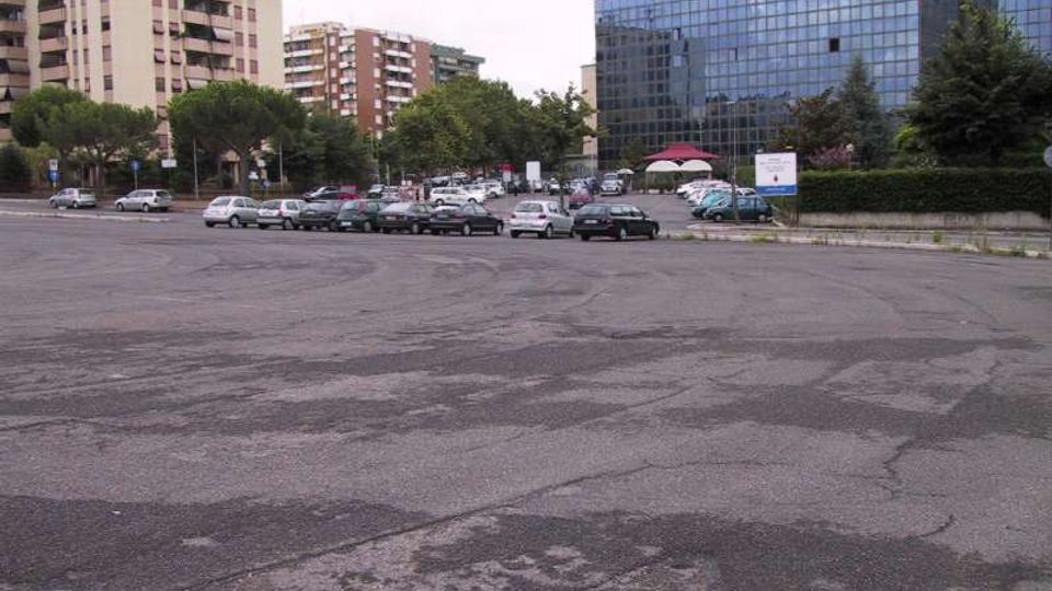 Ampio parcheggio asfaltato con numerose auto in sosta, circondato da alberi e aiuole. Sullo sfondo si vedono palazzi residenziali e un grande edificio moderno con facciata in vetro riflettente.
