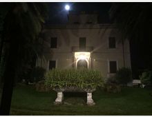 Facciata di una villa a due piani vista di notte, con ingresso ad arco illuminato al centro e finestre simmetriche. Davanti si trova un giardino con prato, un’aiuola rialzata e palme ai lati.
