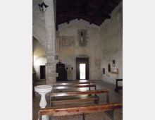 Interno di una piccola chiesa in pietra con soffitto in legno a capriate, panche in legno allineate e fonte battesimale in primo piano. Sul fondo si vedono una porta aperta, una stretta finestra e pareti intonacate con tracce di affreschi.