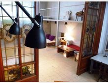 Interno di un loft con soffitti alti e soppalco con scaffalature bianche, affacciato su una zona giorno con divani e tavolino basso. In primo piano si vedono grandi lampade nere e una porta in legno con vetrate decorate che separa l’ambiente.