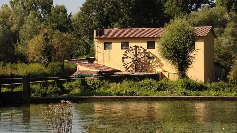 Edificio rurale giallo con tetto in tegole rosse, affacciato su un canale e circondato da vegetazione fitta. Sulla facciata è visibile una grande ruota idraulica in legno, con alberi alti sullo sfondo.