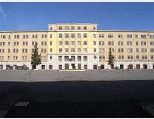 Ampio edificio istituzionale a più piani con facciata giallo chiaro e numerose finestre disposte in griglia, con ingresso centrale rialzato. Davanti si estende una grande piazza asfaltata quasi vuota, con pochi alberi ai lati sotto un cielo sereno.