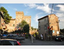 Piazza di un borgo con castello in pietra e torre merlata con orologio sullo sfondo, affiancato da edifici storici intonacati. In primo piano ci sono auto parcheggiate e un passaggio pedonale tra i fabbricati, con alberi e cielo parzialmente nuvoloso.
