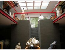 Interno di uno studio d’arte su due livelli con travi rosse a vista, grande lucernario a griglia sul soffitto e scala centrale tra pareti scure. Ai lati ci sono soppalchi con scaffali e materiali, e sul piano inferiore sono visibili sculture e oggetti artistici.