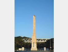 Obelisco in pietra chiara con incisioni verticali, collocato al centro di una piazza aperta. Sullo sfondo si vedono un grande stadio con struttura bianca e alberi, sotto un cielo sereno.