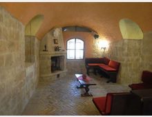 Salotto rustico in pietra con soffitto a volta in mattoni, pavimento in mattoni a spina di pesce e camino in muratura. Al centro c’è un tavolino basso, con panche e divani imbottiti rossi lungo le pareti e una finestra ad arco sul fondo.