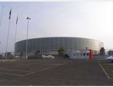 Grande edificio moderno di forma ovale con facciata in vetro e pannelli metallici, recintato e affacciato su un ampio parcheggio asfaltato con segnaletica a terra. In primo piano si vedono un piccolo ingresso con insegna e alcune aste portabandiera e lampioni.