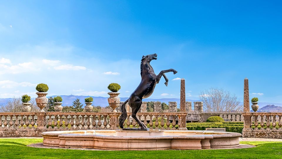 Fontana circolare in pietra con statua in bronzo di un cavallo impennato al centro di un giardino formale. Sullo sfondo si vede una balaustra in pietra con vasi ornamentali e, oltre, colline lontane sotto un cielo azzurro.