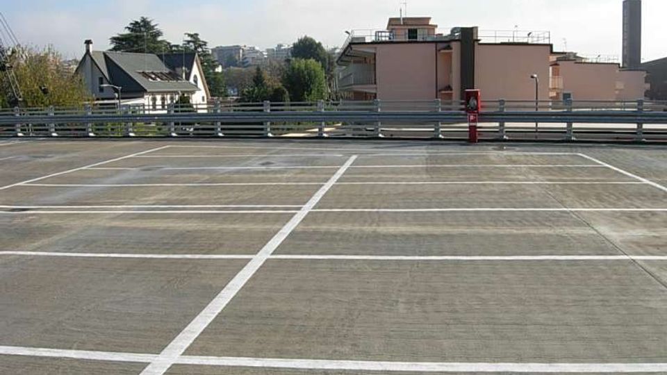 Parcheggio multipiano all’aperto con pavimentazione in cemento e linee bianche che delimitano gli stalli. Sullo sfondo si vedono ringhiere metalliche, edifici residenziali e alberi sotto un cielo parzialmente nuvoloso.