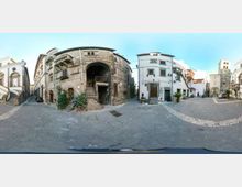 Piazza lastricata in un centro storico, circondata da edifici in pietra e intonaco con archi, scale esterne e balconi. Sullo sfondo si vede una torre campanaria e stradine laterali che si aprono tra le facciate.