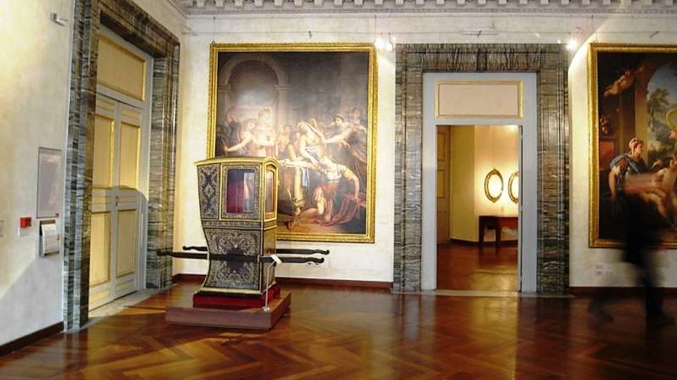 Sala museale con pavimento in parquet lucido, grandi dipinti incorniciati alle pareti e una portantina storica decorata esposta al centro. Porte e passaggi con cornici in marmo conducono a un’altra stanza illuminata.