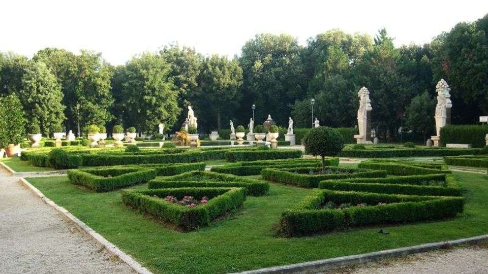 Giardino formale con siepi basse potate a disegni geometrici e vialetti in ghiaia, con ampi prati. Sullo sfondo si vedono statue e vasi ornamentali in pietra davanti a una fitta fila di alberi.
