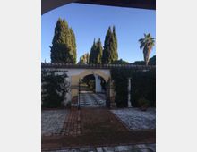 Ingresso di un giardino con cancello in ferro e arco in muratura su un muro bianco con copertura in coppi. Un vialetto pavimentato conduce all’interno, circondato da siepi e alti cipressi, con una palma sullo sfondo sotto un cielo sereno.