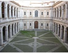 Cortile interno di un edificio storico in pietra, circondato su tre lati da logge a due piani con arcate e colonne. Al centro si vede una pavimentazione a disegno geometrico con fasce di ciottoli e un ampio portale in legno sulla parete di fondo.