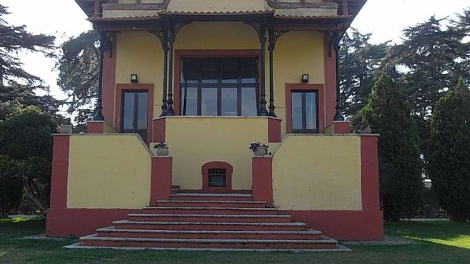 Edificio a due piani in stile villa, con facciata gialla e dettagli rosso mattone, portico centrale con colonne decorative e grande scalinata frontale. La struttura è circondata da un prato e da alberi alti sullo sfondo.