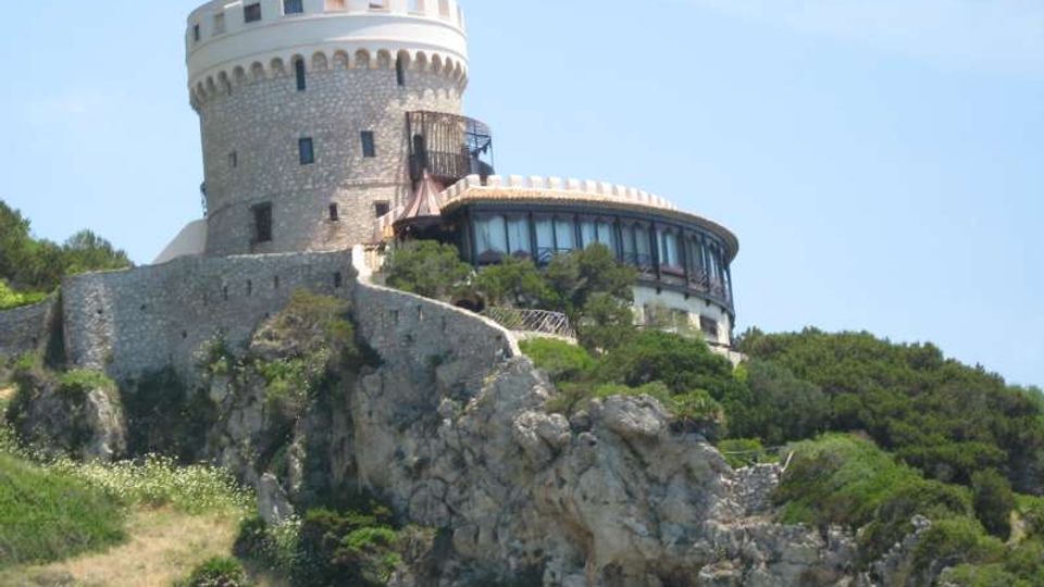 Torre fortificata in pietra con merlature, situata su un promontorio roccioso e collegata a un edificio semicircolare con grandi vetrate. Intorno si vede vegetazione mediterranea su pendii e scogli, sotto un cielo sereno.