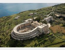 Veduta aerea di un grande complesso edilizio semicircolare in muratura, con cortile interno e ali laterali, situato su una collina verde vicino alla costa. Intorno si vedono strade sterrate, campi terrazzati e pochi edifici isolati, con il mare sullo sfondo.