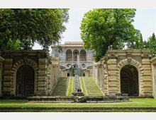 Giardino formale con una lunga scalinata in pietra e un canale d’acqua centrale che sale verso una villa storica con loggia ad archi. Ai lati della scalinata ci sono due grandi portali in muratura decorata e alberi ad alto fusto che incorniciano la scena.