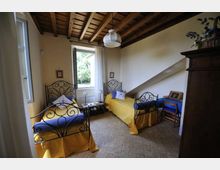 Camera da letto con due letti singoli in ferro battuto, copriletti gialli e cuscini blu, con finestra centrale e piccolo tavolino tra i letti. Soffitto in legno a travi a vista, pavimento in piastrelle e arredi essenziali tra cui una scrivania e un armadio scuro.