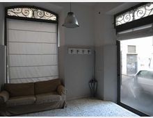 Interno di un piccolo locale con pavimento in graniglia, pareti bicolore e lampada a sospensione. A sinistra c’è un divano sotto una finestra con tendina e grata decorativa, mentre a destra una grande vetrata/porta su strada con tenda e ombrello nell’angolo.