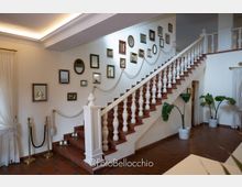 Interno di una hall con grande scala in legno e ringhiera bianca, parete decorata con specchi e cornici di varie dimensioni lungo la salita. Pavimento in cotto, tende bianche alle finestre e piante in vaso ai lati della scala.