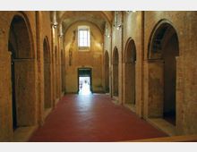 Interno di una sala in mattoni con navata centrale e serie di arcate laterali su colonne, pavimento in cotto rosso e soffitto a volta. In fondo si vedono una finestra alta e una porta aperta che dà verso l’esterno, con illuminazione a faretti lungo le pareti.