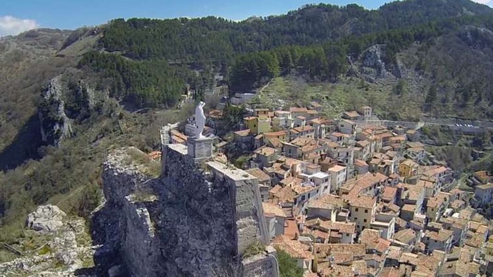 Veduta aerea di un borgo montano con case addossate dai tetti in tegole e strade strette, ai piedi di una rupe rocciosa. In primo piano, una fortificazione in pietra con statua sulla sommità domina il paese, circondato da pendii boscosi e montagne sullo sfondo.