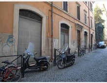 Strada stretta in ciottoli affiancata da un edificio in intonaco color ocra con portoni e finestre ad arco, alcuni con inferriate. Lungo il marciapiede sono parcheggiati scooter e biciclette, con paletti e catene di protezione e un’auto sullo sfondo.