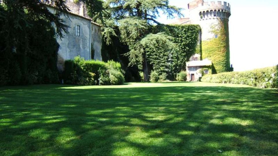 Ampio prato verde in un giardino alberato, con grandi conifere e siepi che delimitano l’area. Sullo sfondo si vedono edifici in pietra, tra cui una torre cilindrica merlata parzialmente coperta da edera.