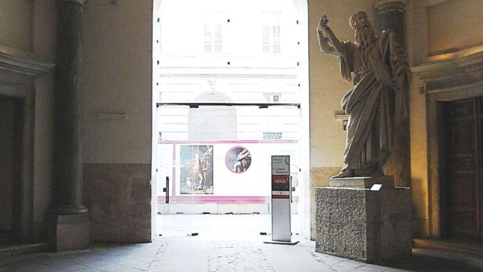 Interno di un portico monumentale con pavimento in pietra e una corsia centrale in ciottoli che conduce a un grande arco d’ingresso illuminato verso l’esterno. A destra si trova una grande statua su piedistallo accanto a colonne e pareti con modanature classiche.