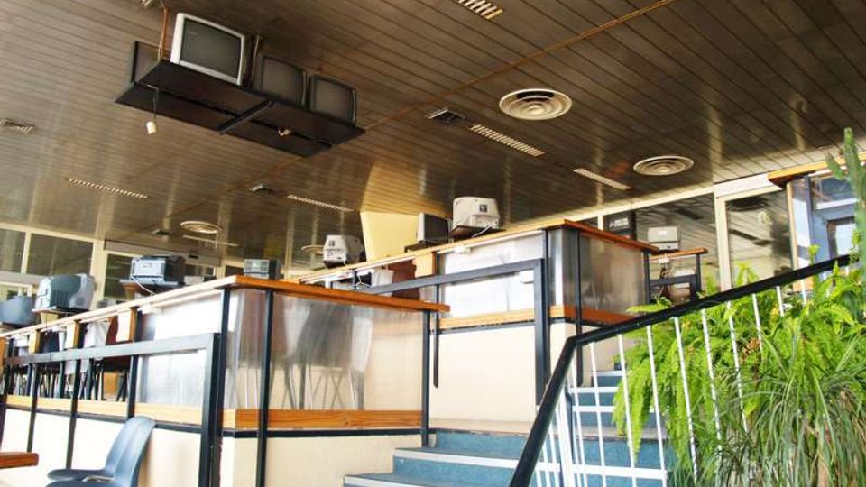Interno di un ufficio open space su due livelli con scale e corrimano in metallo, postazioni con computer e grandi vetrate lungo il perimetro. Soffitto a pannelli con luci fluorescenti, diffusori d’aria e vecchi monitor sospesi, con piante ornamentali vicino alla scala.