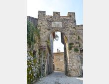 Antica porta urbana in pietra con arco a tutto sesto e merlature superiori, con targa e lanterna sospesa al centro dell’apertura. Sotto l’arco passa una stretta strada acciottolata tra edifici in muratura e pareti con vegetazione rampicante.